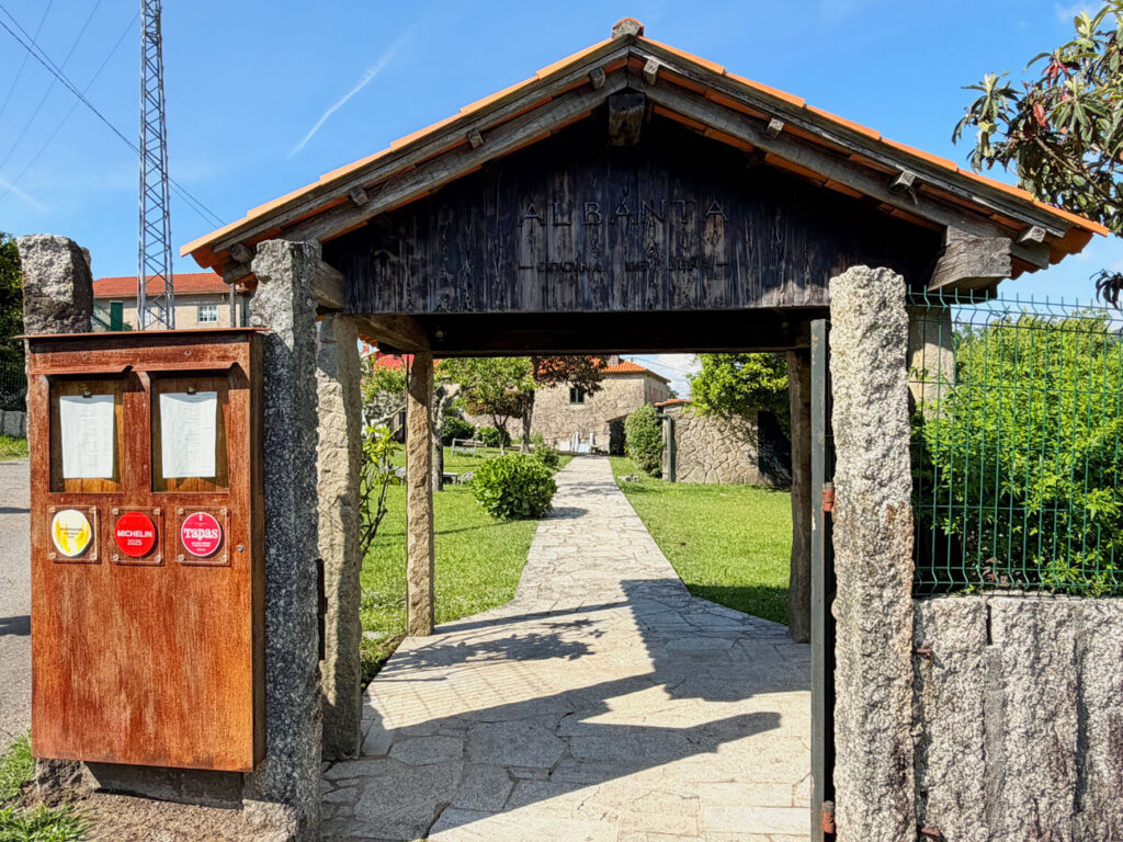 Entrada exterior del restaurante Albanta Cocina de Leña en Pontevedra, con portalón de madera y granito, rótulo del restaurante y distintivos Michelin 2025 y Tapas en el tablón de menús.