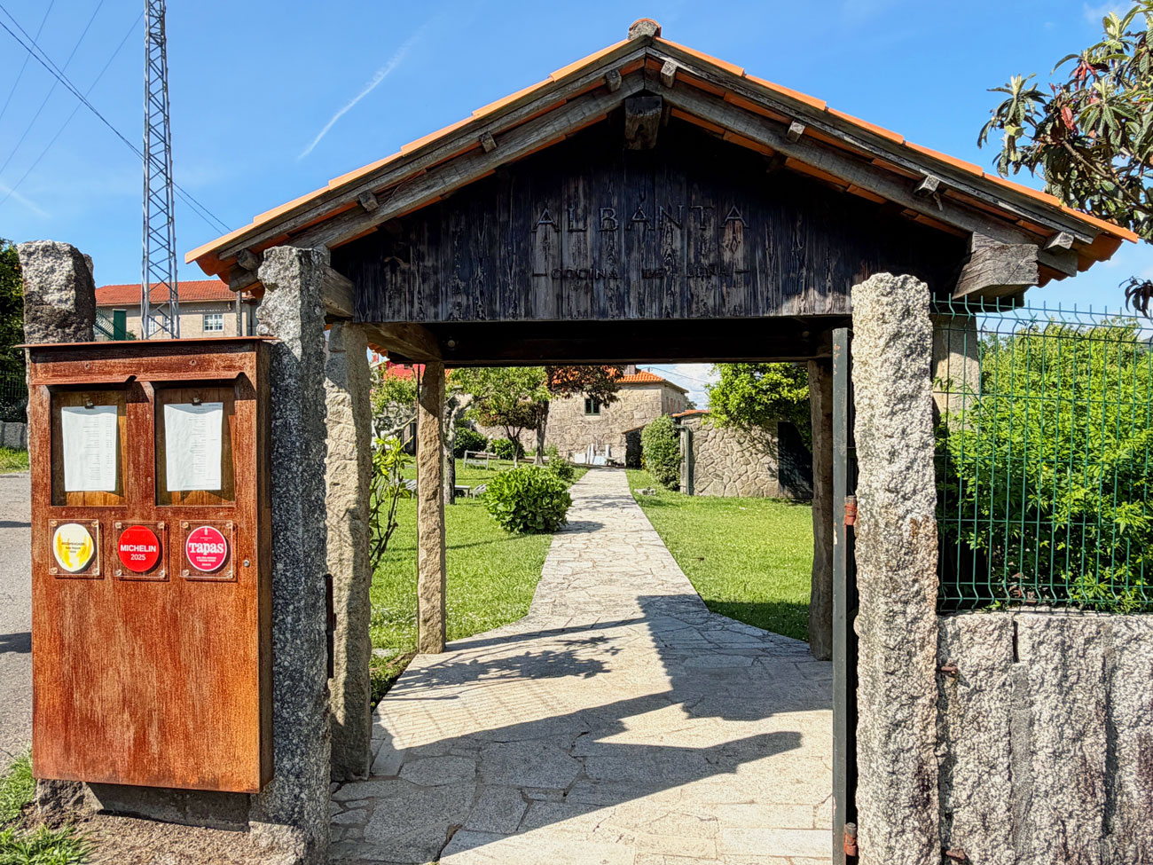 Entrada exterior del restaurante Albanta Cocina de Leña en Pontevedra, con portalón de madera y granito, rótulo del restaurante y distintivos Michelin 2025 y Tapas en el tablón de menús.